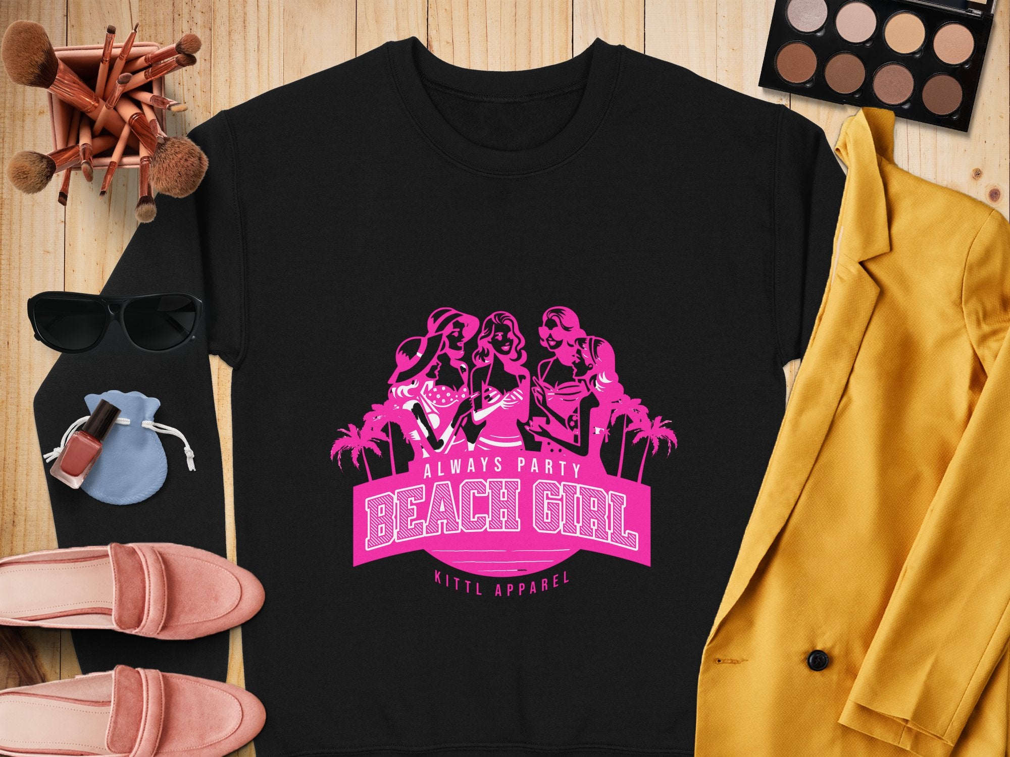 Flat lay of a Garment Graphics black sweatshirt with a pink Always Party Beach Girl design, showcasing three women and palm trees. Its surrounded by a gold jacket, makeup palette, sunglasses, shoes, perfume, and brushes on a wooden background.