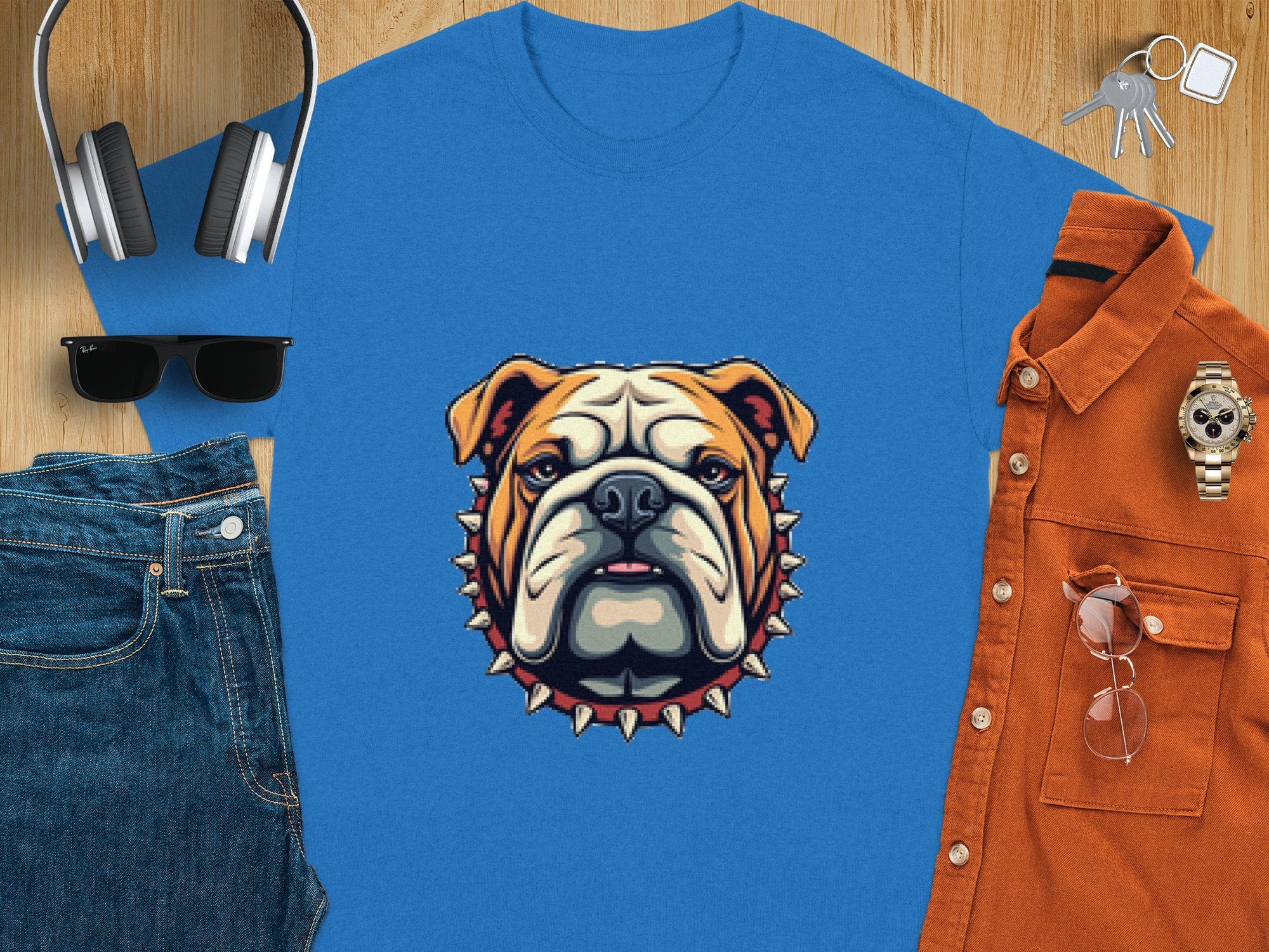 A Garment Graphics blue T-shirt with a bulldog in a spiked collar is on a wooden surface, surrounded by headphones, sunglasses, a brown shirt, watch, keys, and jeans—ideal for those who love stylish details and family sizing options.