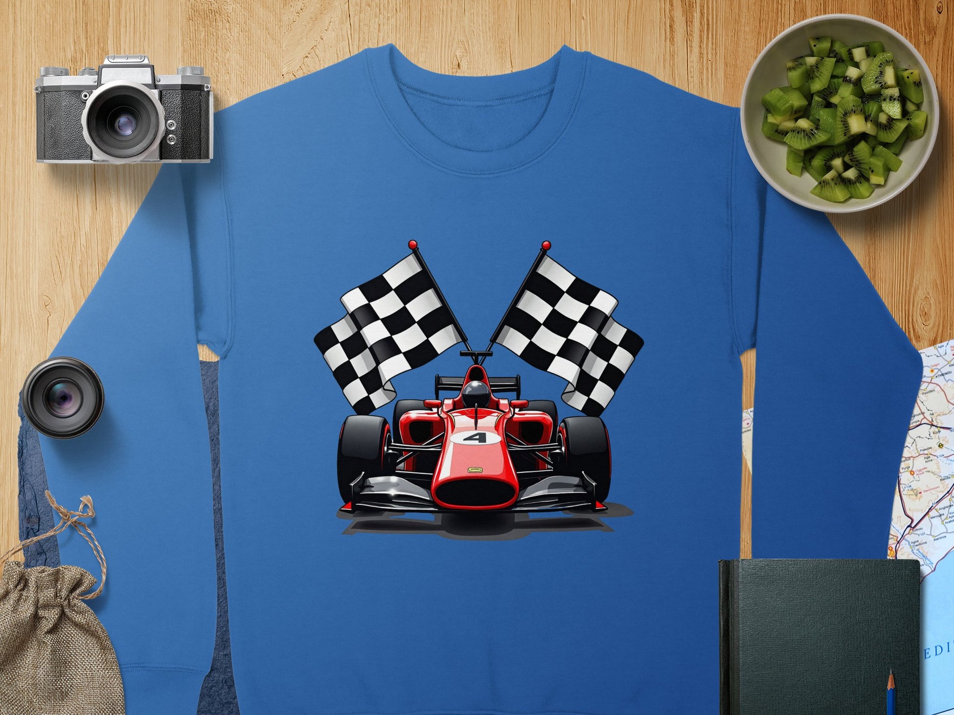 A vibrant blue Garment Graphics sweatshirt featuring a red racing car and checkered flags is displayed on a wooden surface alongside a camera, lens, chopped kiwis, a bag, and a black notebook on a map—perfect for motorsport enthusiasts.