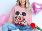 A person sits holding a red rose and wearing a Garment Graphics pink unisex sweatshirt featuring a cartoon donut character. Pink heart-shaped balloons float behind them. They smile in ripped jeans, though their face is partially out of frame.