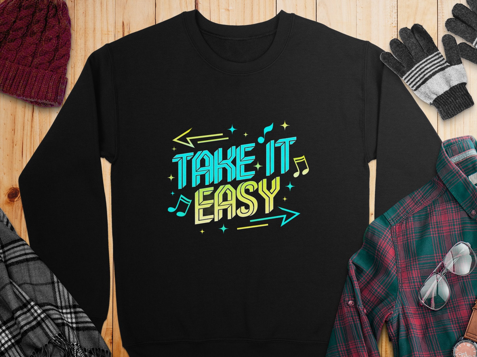 On a wooden surface lies a black unisex sweatshirt by Garment Graphics, featuring TAKE IT EASY in bold, colorful letters with musical notes and arrows. Nearby are casual essentials: a red knit hat, gloves, a plaid shirt, and a scarf.
