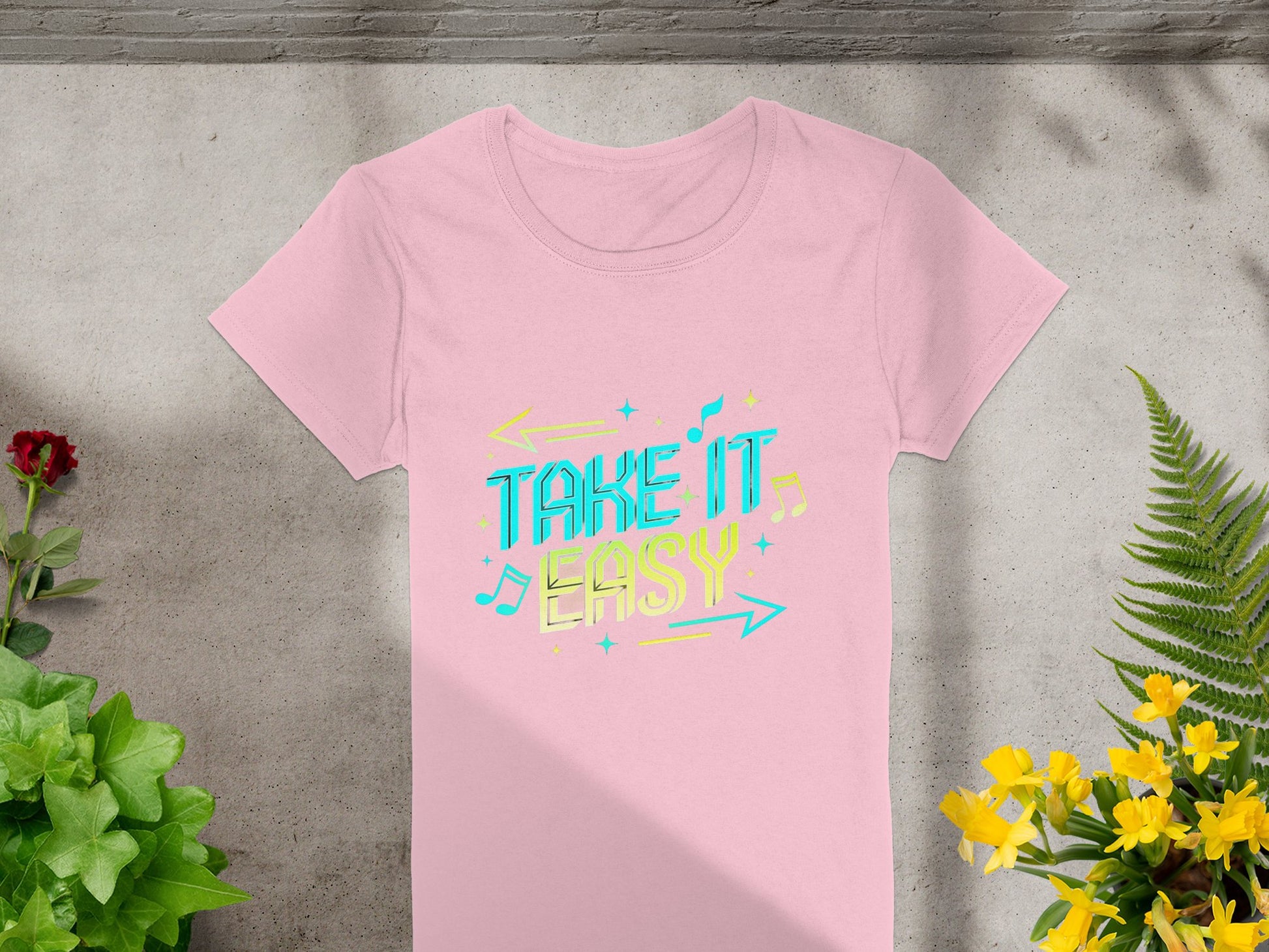 A pink Take It Easy T-shirt from Garment Graphics lies on a concrete surface, featuring vibrant graphics of colorful letters with music notes and arrows. Its stylishly complemented by green plants and yellow flowers around it.