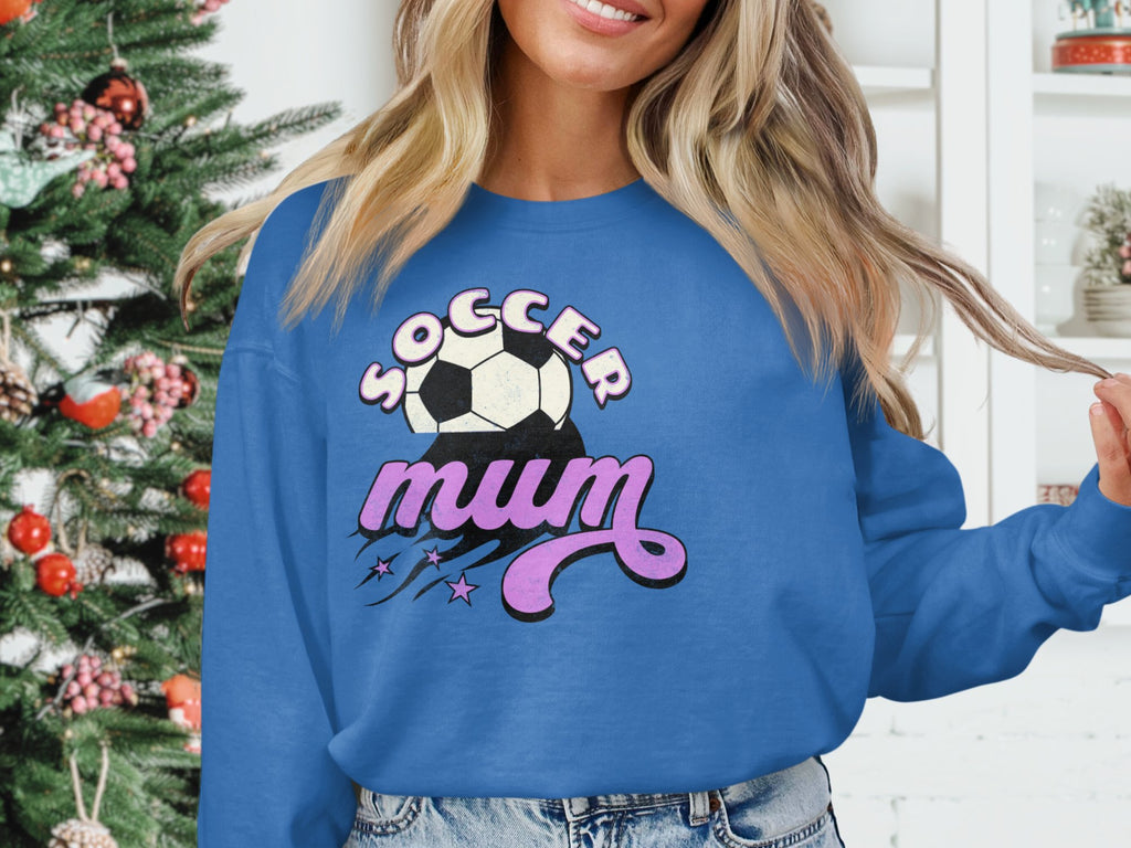 A woman poses indoors before a festive Christmas tree and white cabinet, wearing a blue Soccer Mum sweatshirt with a soccer ball motif and decorative text by Garment Graphics.