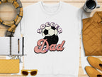A Garment Graphics retro white t-shirt with Soccer Dad text and soccer ball graphic is displayed on wood, surrounded by a scarf, coffee mug, notebook, pen, jacket, sunglasses, and quilted clutch in warm yellow and brown hues.