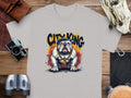 The Garment Graphics gray t-shirt, featuring a bulldog with a crown and gold chain, sits on wood against an orange-black graffiti background reading City King. It exudes unique style with surrounding accessories like a camera, leather gloves, and binoculars.