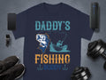 A flat lay showcases a navy Graphic Print T-Shirt from Garment Graphics, labeled Daddys Fishing Buddy with a fish and boat illustration. Ideal for fishing fans, its paired with black sneakers, a smartwatch, a smartphone, a portable speaker, and two dumbbells.