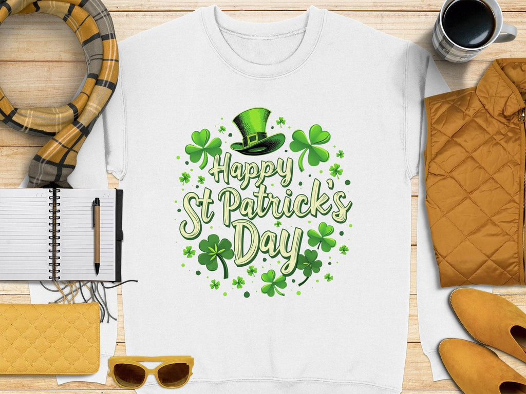 The Garment Graphics white T-shirt featuring Happy St. Patricks Day in green, surrounded by shamrocks and a leprechaun hat, adds festive cheer on a wooden surface with accessories like a scarf, pen, notebook, yellow clutch, sunglasses, and coffee nearby.