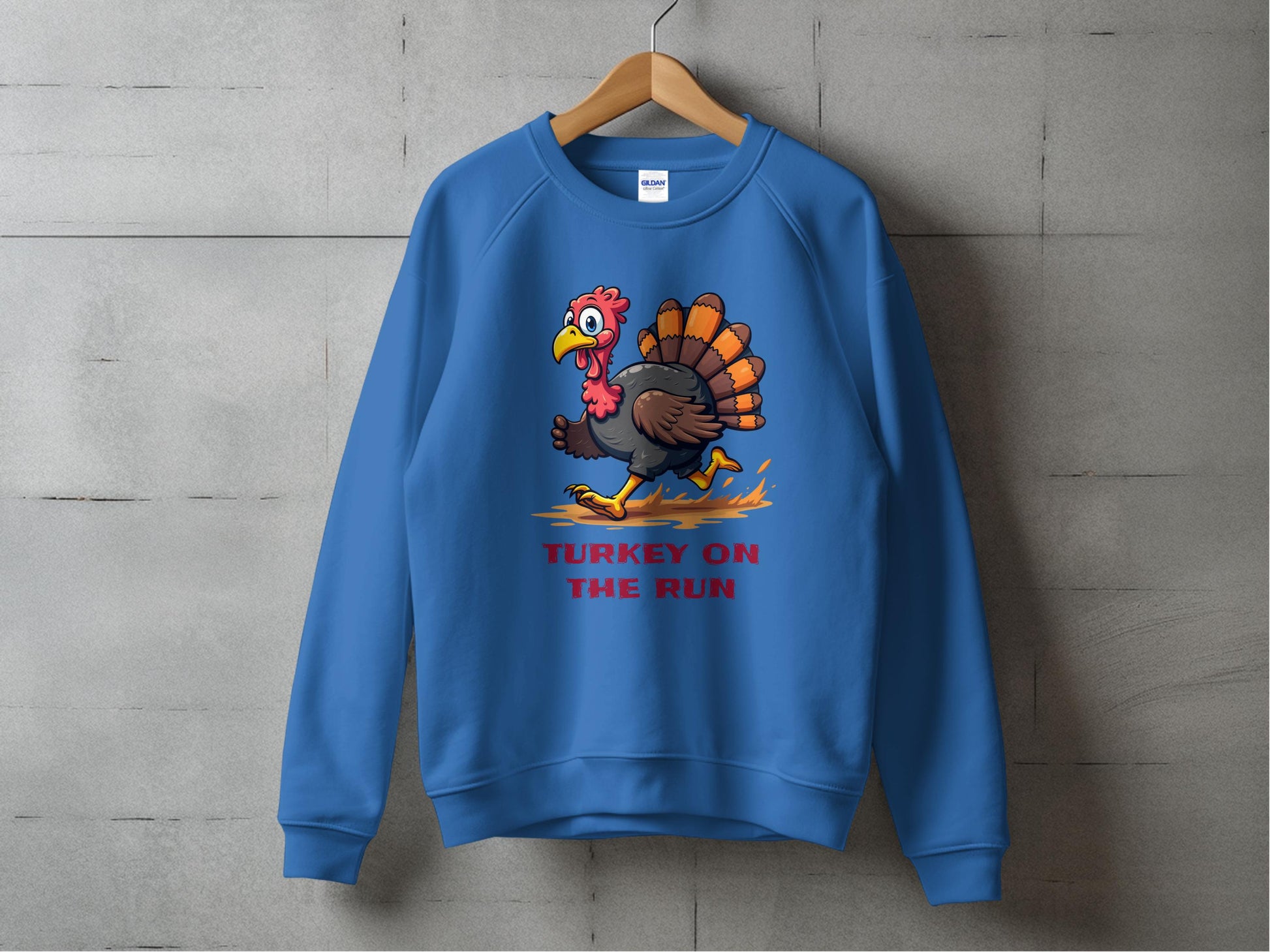 A blue sweatshirt by Garment Graphics hangs on a wooden hanger against a gray wall, showcasing a humorous cartoon turkey with TURKEY ON THE RUN in red letters, highlighting their collection of funny graphic sweatshirts in unisex sizing.