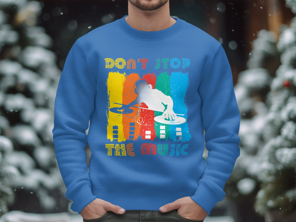 A person confidently wears a blue Garment Graphics DJ sweatshirt, featuring a DJ silhouette on turntables with the words DONT STOP THE MUSIC in colorful letters set against snowy trees. Available in unisex sizing for music lovers seeking comfort and style.