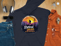 A flat lay features Garment Graphics navy hiking hoodie with a sunset mountain design and Retired 2025, Not My Problem Anymore text, paired with jeans, an orange shirt, wristwatch, sunglasses, headphones, and keys on a wooden surface.