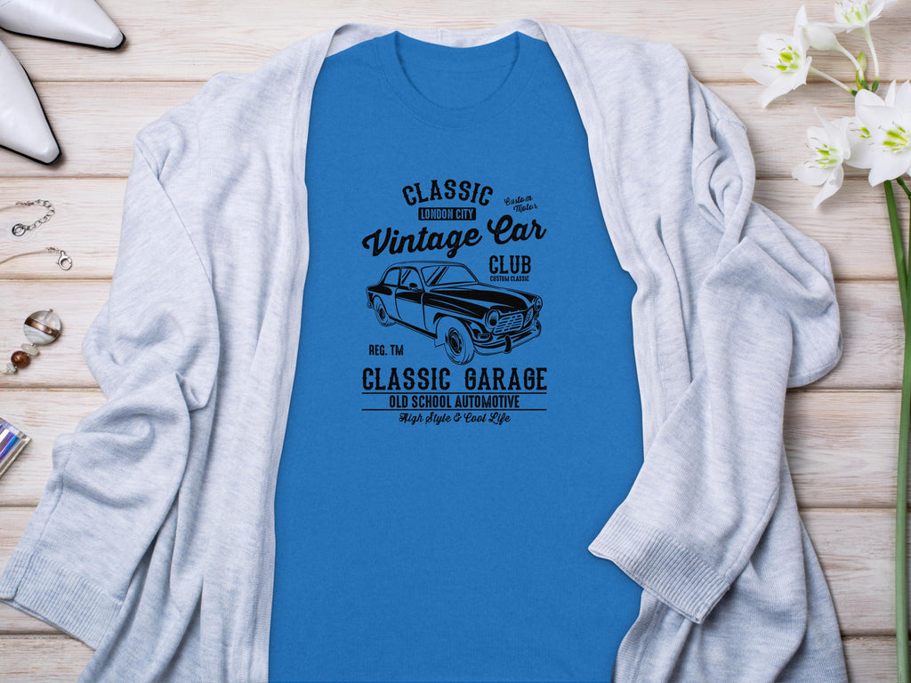 Garment Graphics’ blue T-shirt, featuring a vintage car and Vintage Car Club, Classic Garage, Old School Automotive text, showcases retro automotive style. Styled on wood with a gray cardigan, it’s surrounded by flowers, a watch, and earrings for an ideal classic fit look.