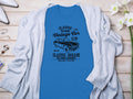Garment Graphics’ blue T-shirt, featuring a vintage car and Vintage Car Club, Classic Garage, Old School Automotive text, showcases retro automotive style. Styled on wood with a gray cardigan, it’s surrounded by flowers, a watch, and earrings for an ideal classic fit look.