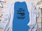 Garment Graphics’ blue T-shirt, featuring a vintage car and Vintage Car Club, Classic Garage, Old School Automotive text, showcases retro automotive style. Styled on wood with a gray cardigan, it’s surrounded by flowers, a watch, and earrings for an ideal classic fit look.