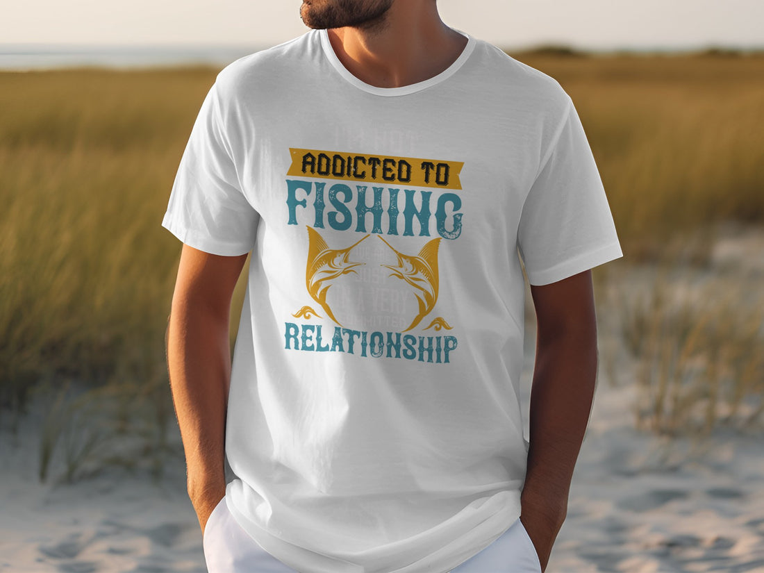 A man stands on sandy ground near tall grass, wearing a white Garment Graphics Fishing T-shirt with Addicted to Fishing and Relationship, featuring two fish. The beachy backdrop perfectly suits this apparel for passionate anglers.