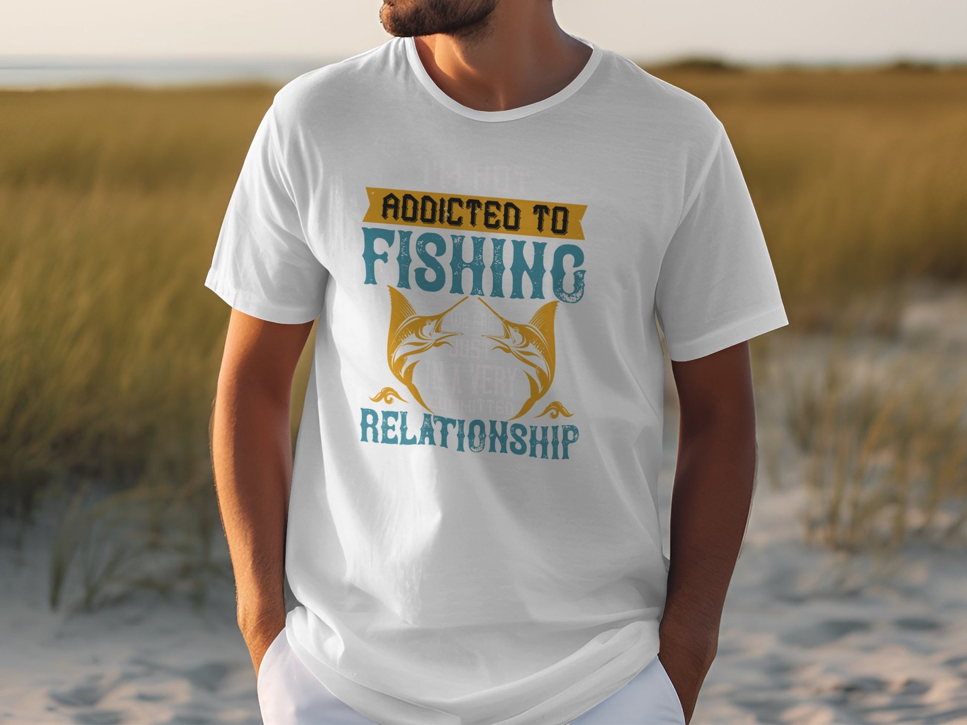 A man stands on sandy ground near tall grass, wearing a white Garment Graphics Fishing T-shirt with Addicted to Fishing and Relationship, featuring two fish. The beachy backdrop perfectly suits this apparel for passionate anglers.