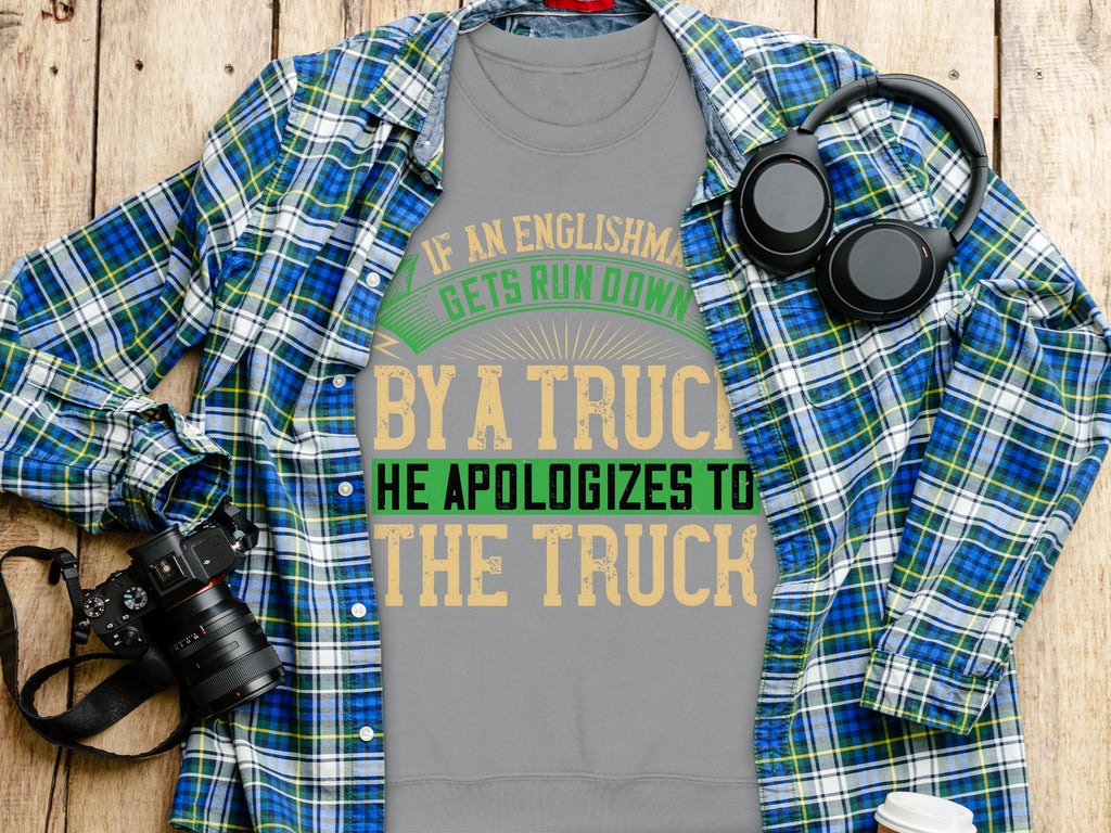 A gray Garment Graphics t-shirt with the witty design If an Englishman gets run down by a truck, he apologizes to the truck lies on a wooden surface. This unisex piece is displayed next to a blue plaid shirt, a black camera, and sleek black headphones.