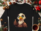 A black shirt from Garment Graphics with a cartoon eagle giving a thumbs-up is laid on wood, surrounded by Christmas decorations like pine branches, pinecones, ornaments, and a cup of dark liquid. The shirt is available in unisex sizes.