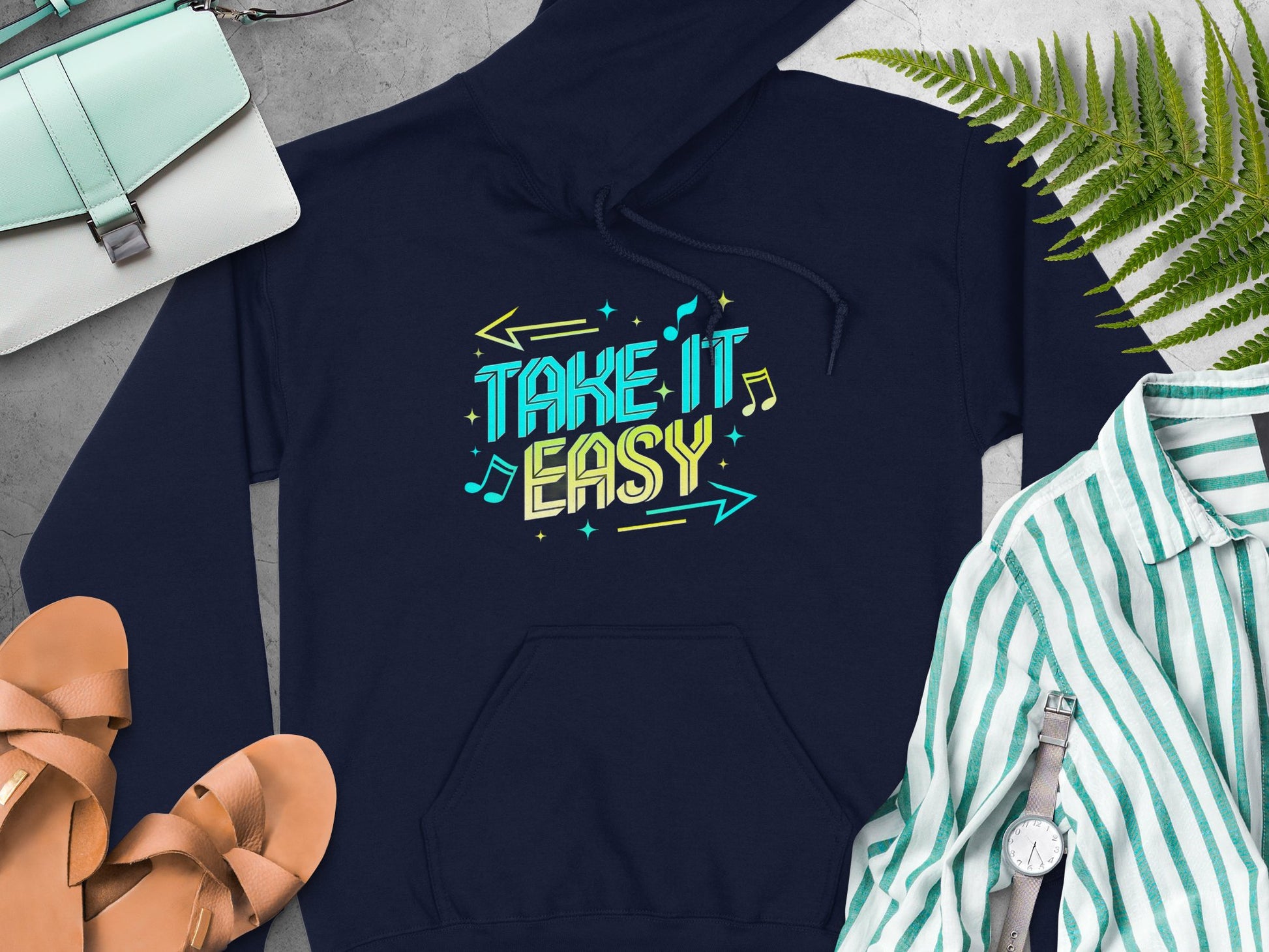 A navy hoodie by Garment Graphics, featuring a vibrant Take It Easy design in bright blue and yellow, is laid on a gray surface surrounded by sandals, a green and white striped shirt, a watch, purse, and fern leaves for the ultimate comfortable fashion vibe.