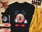 Garment Graphics black unisex sweatshirt features a whimsical donut character with mouse ears, surrounded by makeup brushes, an eyeshadow palette, sunglasses, blush, lipstick, pink loafers, and a yellow blazer on a wooden surface.