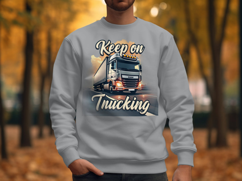 A person wears a Garment Graphics inspirational sweatshirt featuring a large truck and the phrase Keep on Trucking. The backdrop shows an open road with tree-lined paths draped in vivid autumn leaves.