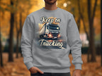 A person wears a Garment Graphics inspirational sweatshirt featuring a large truck and the phrase Keep on Trucking. The backdrop shows an open road with tree-lined paths draped in vivid autumn leaves.