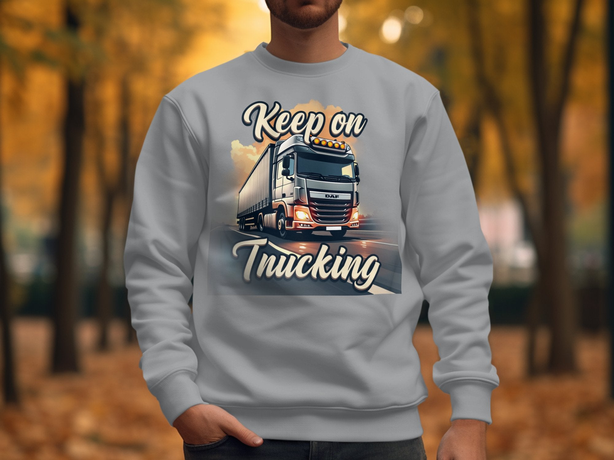 A person wears a Garment Graphics inspirational sweatshirt featuring a large truck and the phrase Keep on Trucking. The backdrop shows an open road with tree-lined paths draped in vivid autumn leaves.