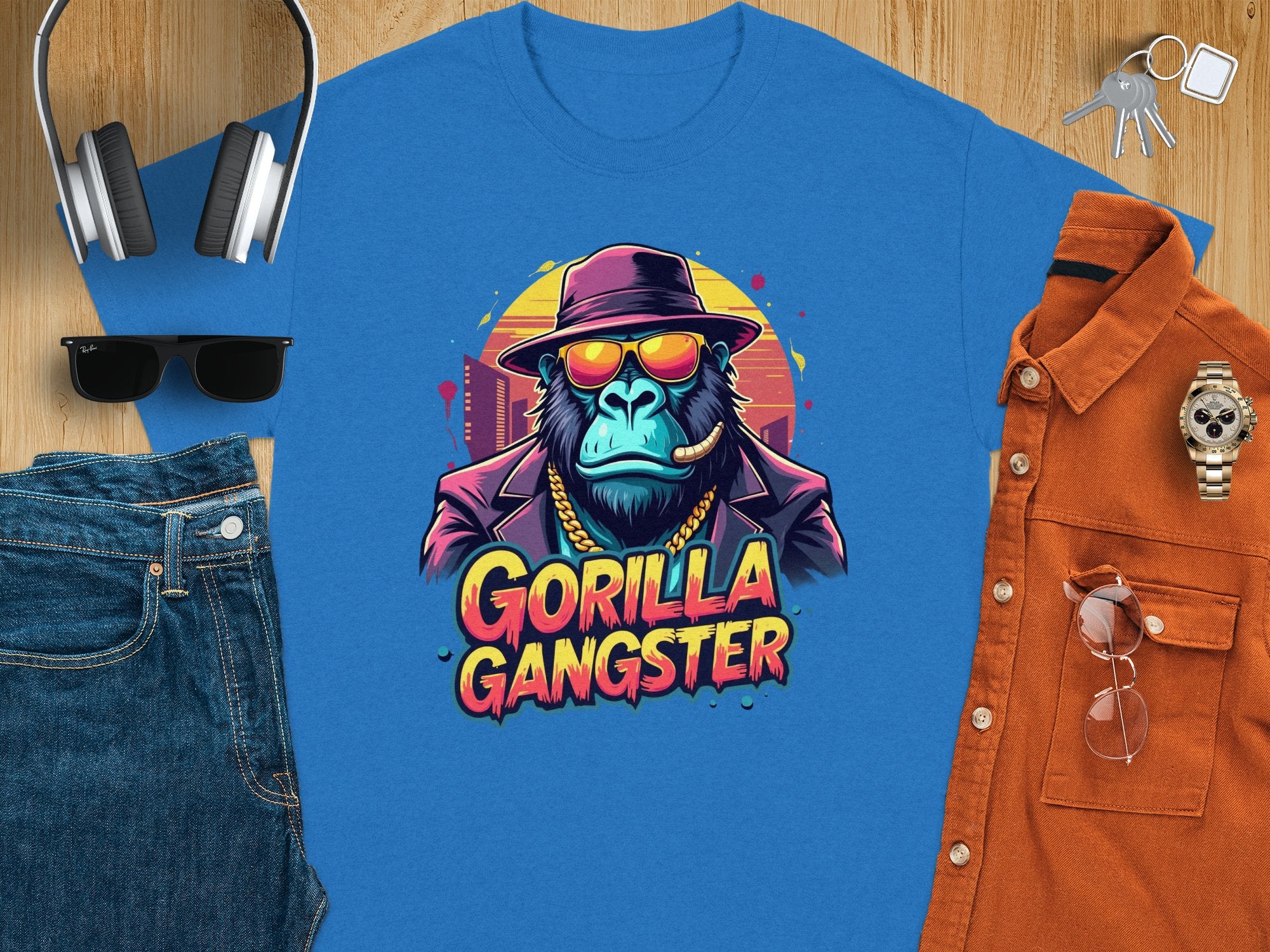 A stylish blue T-shirt by Garment Graphics, featuring a bold Gorilla Gangster design with a hat, sunglasses, and cigar, is paired with jeans, a brown shirt, wristwatch, sunglasses, headphones, and keys on wood. Ideal for those making bold fashion statements with graphic tees.