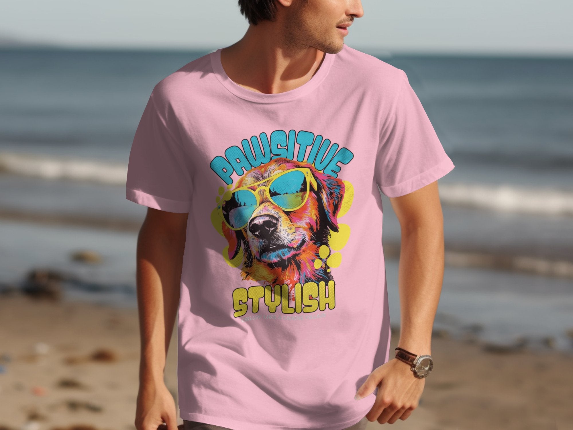Wearing Garment Graphics light pink T-shirt with a vibrant dog-in-sunglasses graphic and Chillin Like A Canine Villain slogan, a person strolls along the sandy beach as the ocean waves gently lap in the background.