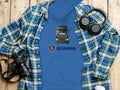 A blue Scania Truck Hoodie from Garment Graphics, with a graphic design, lies on a wooden surface. Its topped with a green-and-blue plaid shirt and surrounded by items for truck enthusiasts like headphones, a camera, and a white cup.