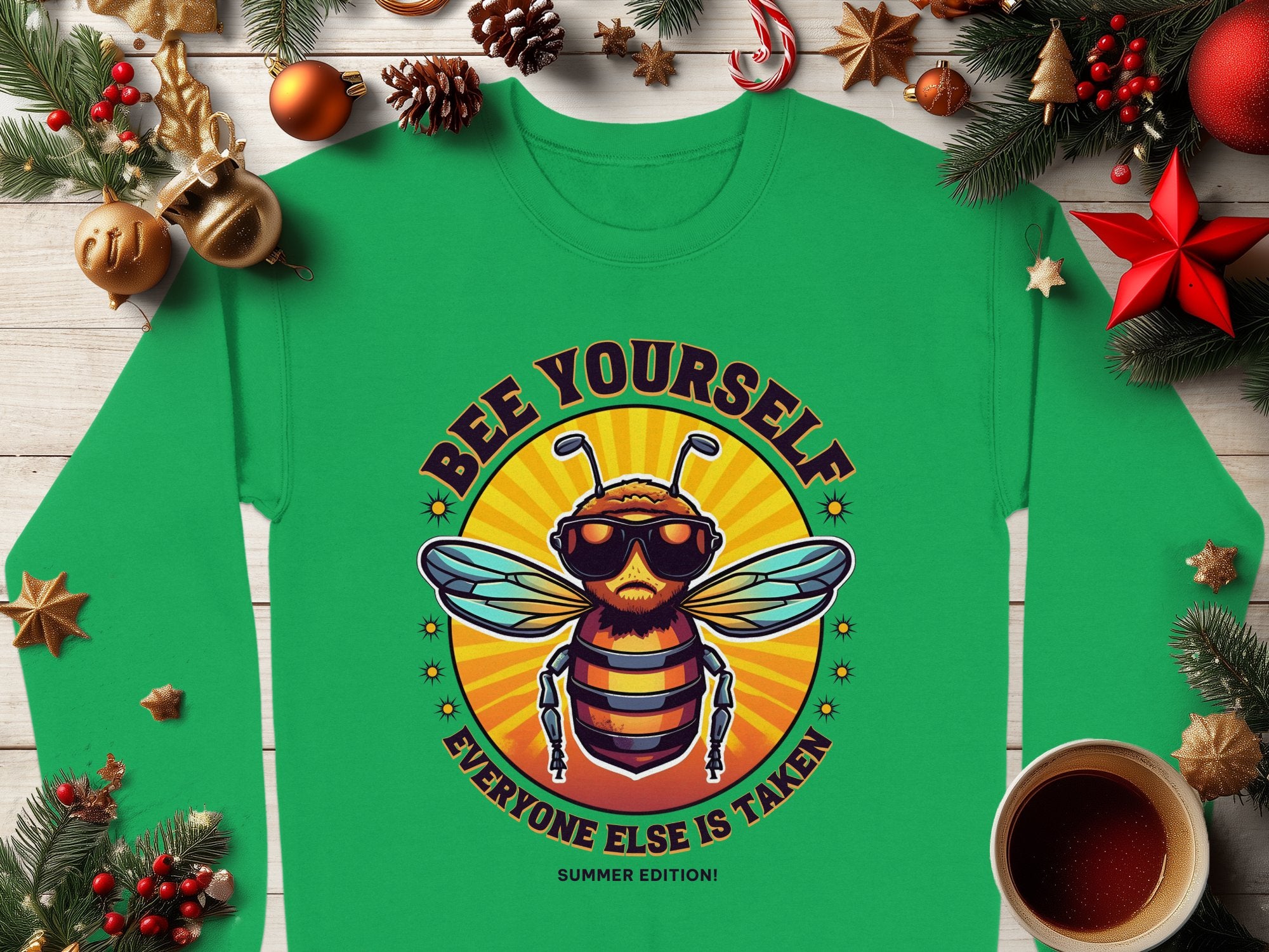 Garment Graphics Summer Edition Sweatshirts collection offers a green unisex sweatshirt featuring a cartoon bee in sunglasses with the motivational text BEE YOURSELF: EVERYONE ELSE IS TAKEN, surrounded by holiday decorations like pine branches, baubles, and a steaming coffee cup.