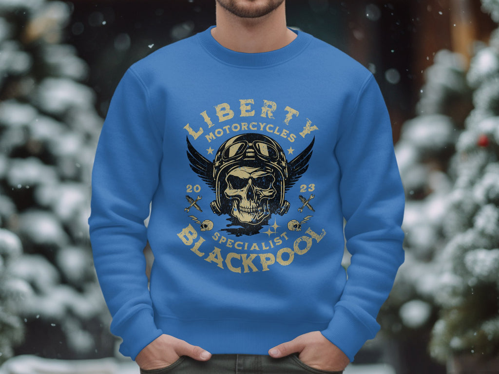 Someone shows off a blue sweatshirt from Garment Graphics Blackpool 2023 collection, featuring a winged skull with goggles and helmet. Bold Liberty Motorcycles Specialist Blackpool 2023 text enhances the design against snowy trees.