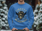 Someone shows off a blue sweatshirt from Garment Graphics Blackpool 2023 collection, featuring a winged skull with goggles and helmet. Bold Liberty Motorcycles Specialist Blackpool 2023 text enhances the design against snowy trees.