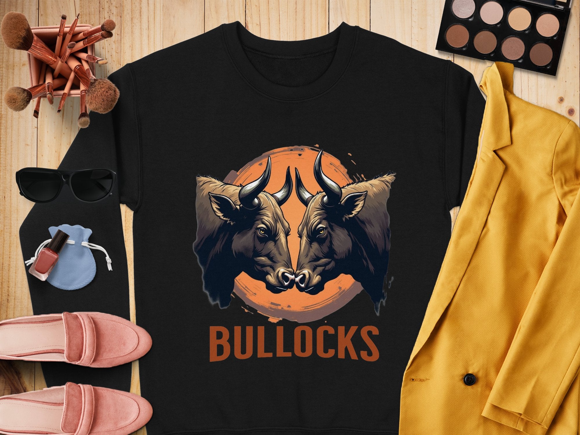 Garment Graphics presents an eye-catching black sweatshirt with a striking bullocks graphic print on a wooden backdrop. Pair it with a yellow jacket, pink shoes, sunglasses, makeup essentials, and a blue drawstring bag for a chic outfit.