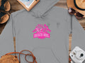 Garment Graphics gray hoodie with vibrant pink graphics of women, palm trees, and Always Party Beach Girl Hotel Apparel text. Styled with a notebook, map, straw hat, sandals, sunglasses, rope bag, and a white instant camera on a wooden surface. Casual style perfection!.