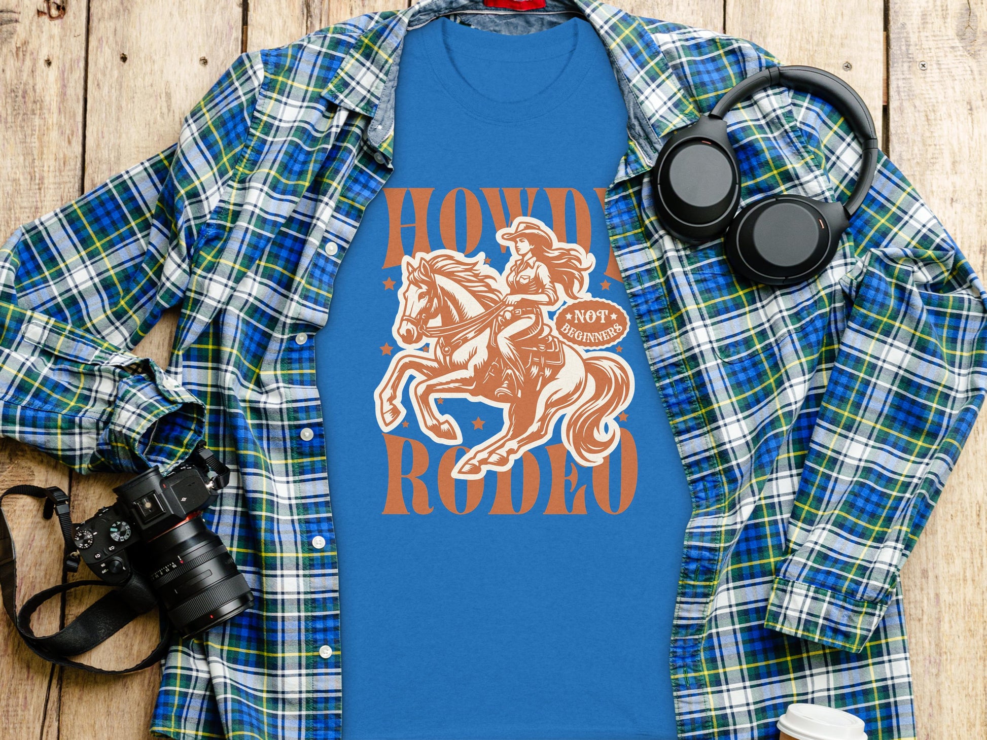 A blue Garment Graphics Rodeo T-Shirt shows a cowboy on horseback with Howdy Rodeo, Not My First Rodeo text. Perfect for enthusiasts, it’s styled on wood surrounded by a plaid shirt, camera, headphones, and cup.