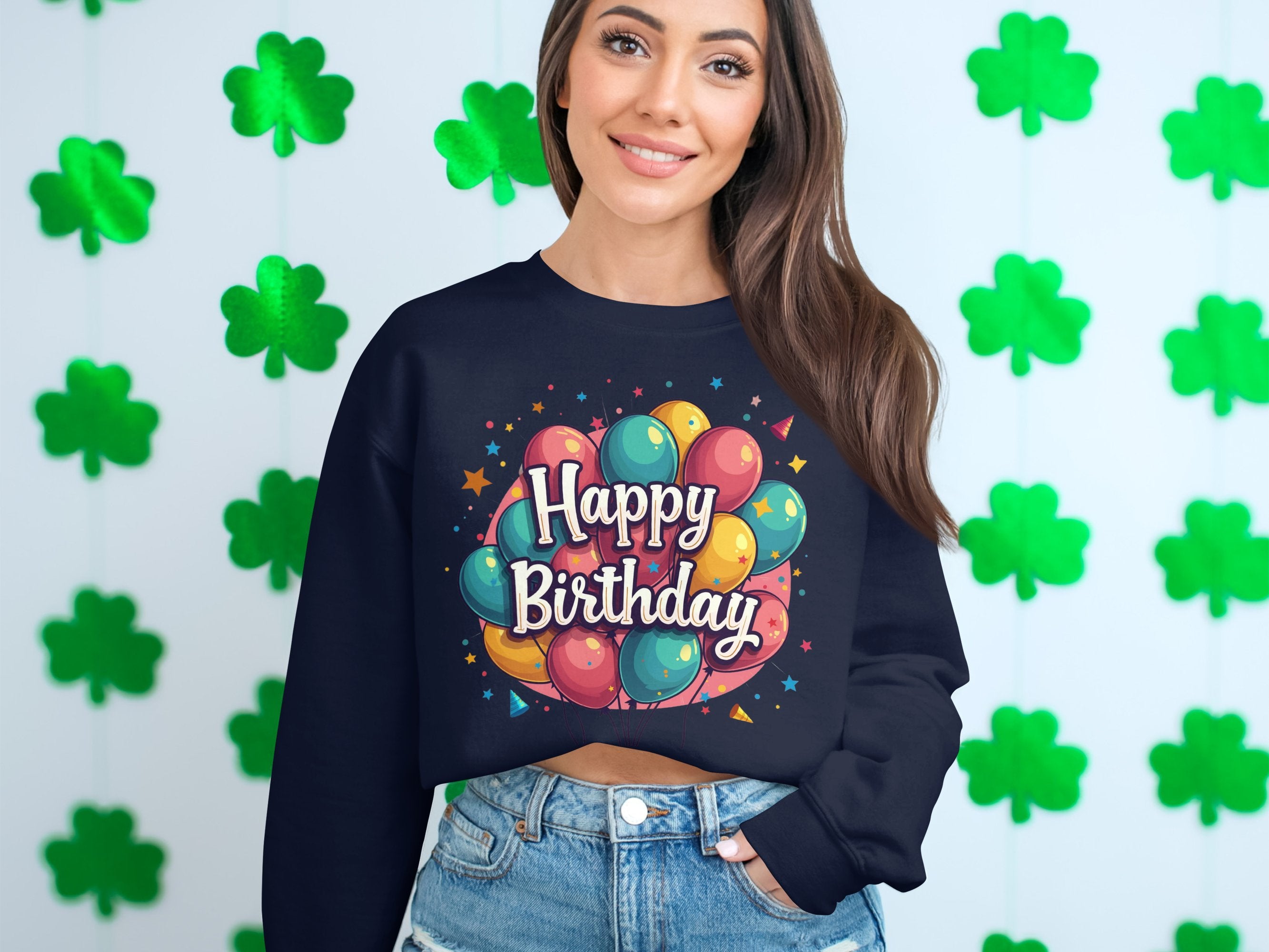A person with long hair celebrates by a green clover backdrop, wearing a Garment Graphics dark Happy Birthday sweatshirt adorned with colorful balloons and denim shorts. The unisex sizing of this festive sweatshirt makes it ideal for any occasion.