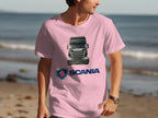 A truck enthusiast wears a pink Garment Graphics T-shirt featuring a Scania truck image and logo. They pose on a tranquil beach with the ocean in the background.