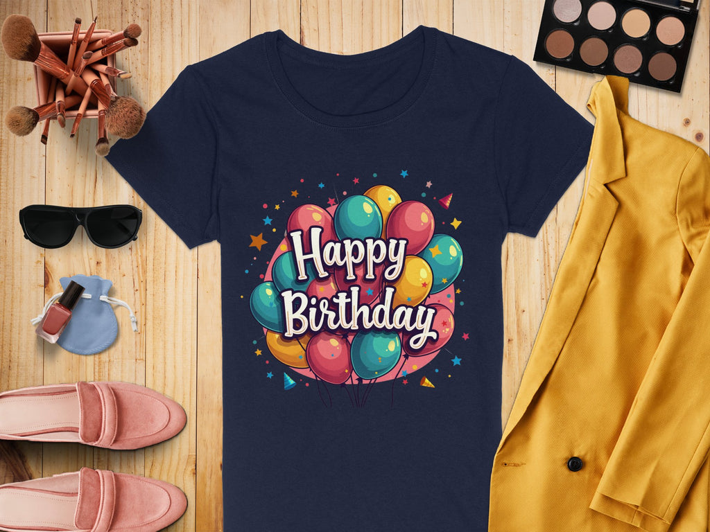 A vibrant Garment Graphics navy blue T-shirt with colorful balloons and Happy Birthday print, crafted from 100% cotton, rests on wood. Nearby are a mustard yellow blazer, makeup items, pink shoes, sunglasses, and a small blue purse.