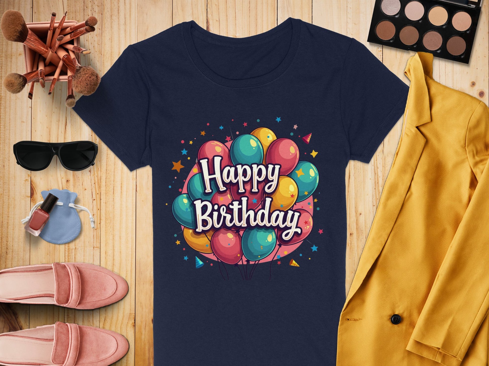 A vibrant Garment Graphics navy blue T-shirt with colorful balloons and Happy Birthday print, crafted from 100% cotton, rests on wood. Nearby are a mustard yellow blazer, makeup items, pink shoes, sunglasses, and a small blue purse.