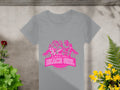 A gray T-shirt from Garment Graphics features a pink graphic of three women, palm trees, and Always Party Beach Girl, with Kitty Apparel below. Its laid out beside vibrant green foliage and yellow flowers.