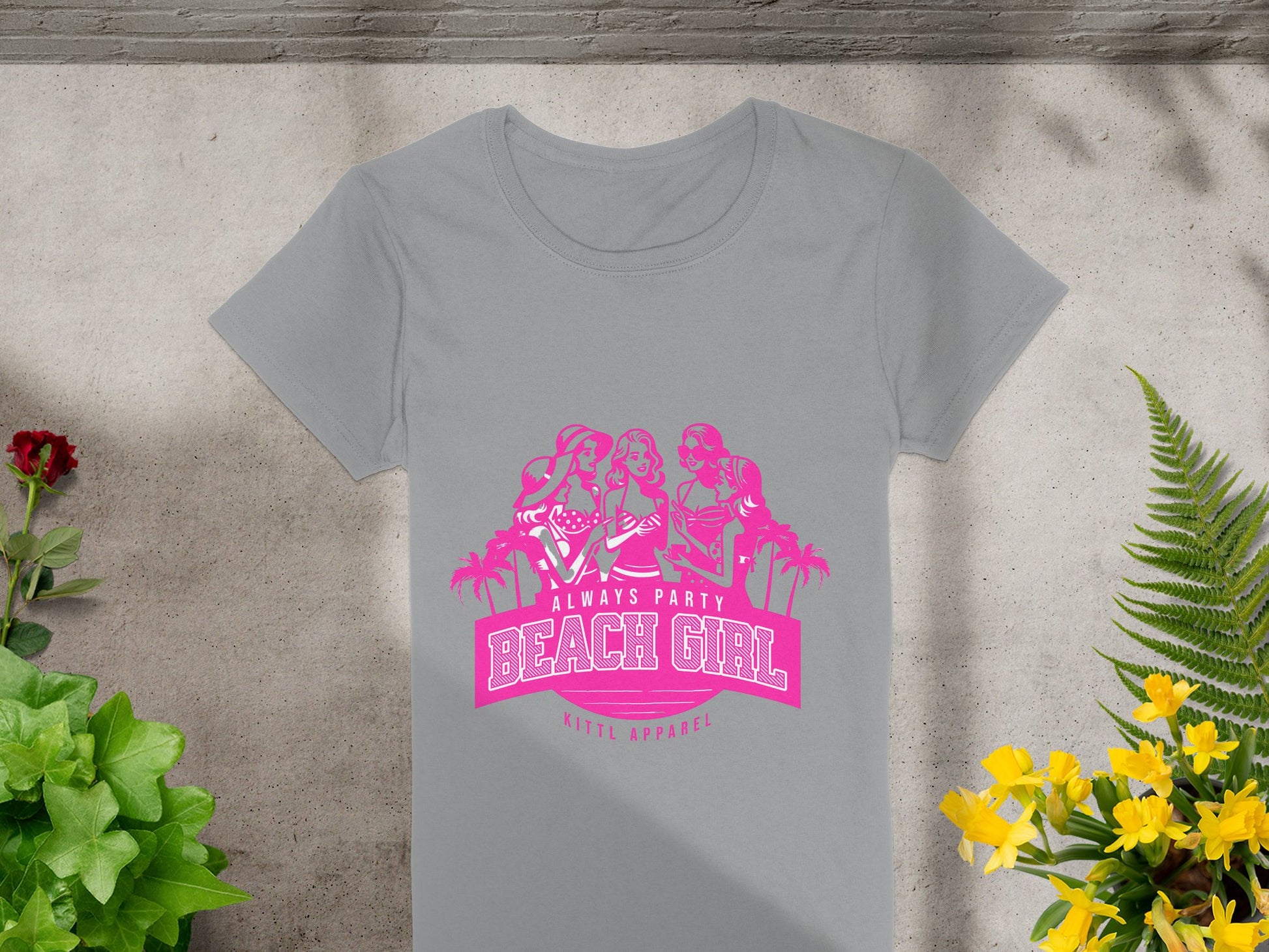 A gray T-shirt from Garment Graphics features a pink graphic of three women, palm trees, and Always Party Beach Girl, with Kitty Apparel below. Its laid out beside vibrant green foliage and yellow flowers.