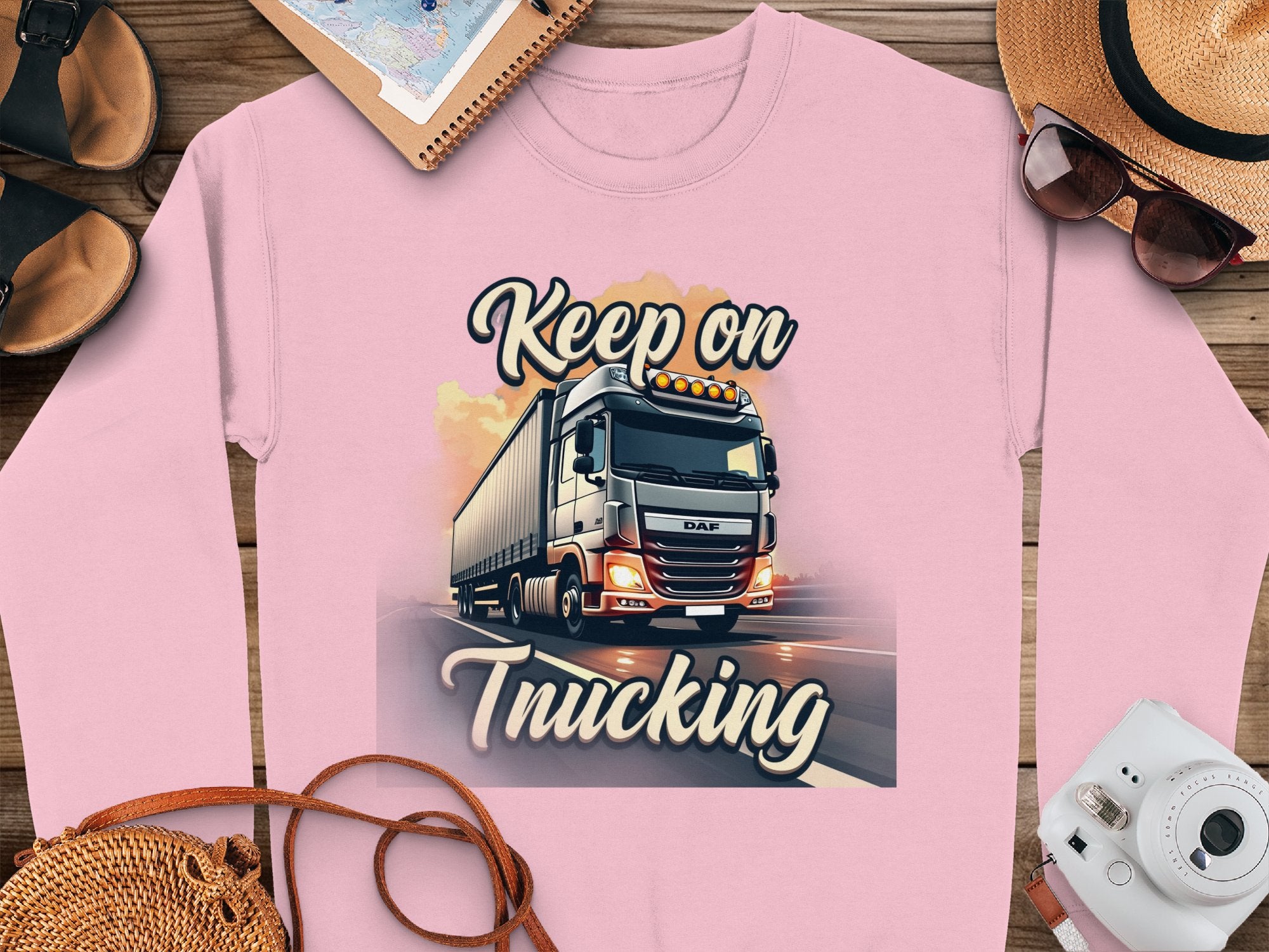 The Garment Graphics pink graphic sweatshirt showcases a vivid truck illustration with the Keep on Trucking motto. Ideal for any style, it matches well with a hat, sunglasses, sandals, and a notebook on a wooden surface. Unisex sizing available.