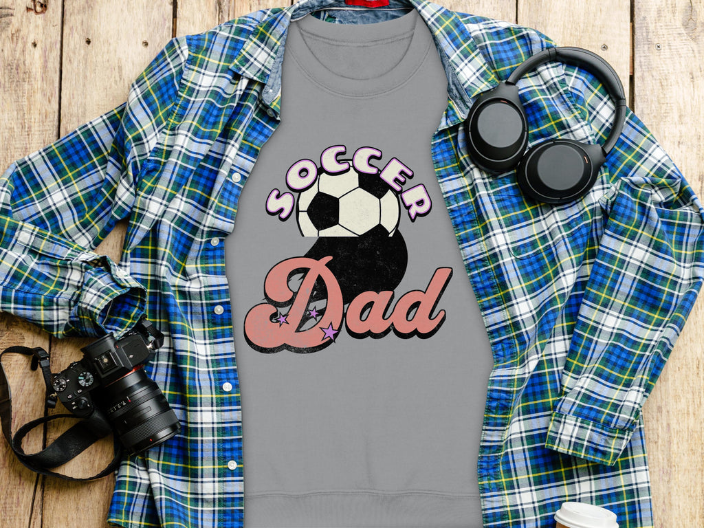 A flat lay showcases a Garment Graphics Soccer Dad sweatshirt featuring a soccer ball design. Nearby are a retro blue plaid shirt, a camera, headphones, and a disposable coffee cup, all arranged on wood.