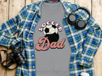 A flat lay showcases a Garment Graphics Soccer Dad sweatshirt featuring a soccer ball design. Nearby are a retro blue plaid shirt, a camera, headphones, and a disposable coffee cup, all arranged on wood.