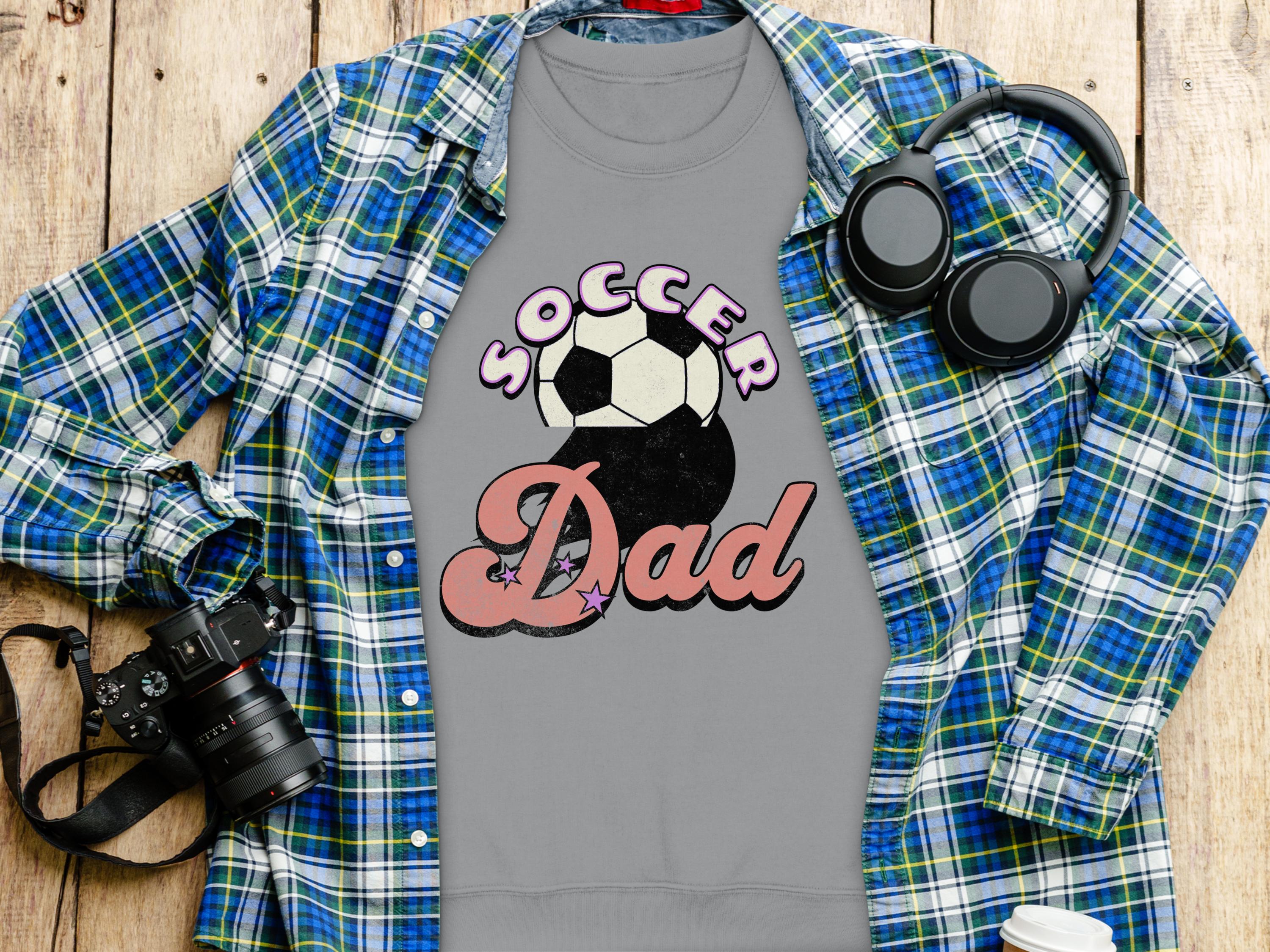 A flat lay showcases a Garment Graphics Soccer Dad sweatshirt featuring a soccer ball design. Nearby are a retro blue plaid shirt, a camera, headphones, and a disposable coffee cup, all arranged on wood.