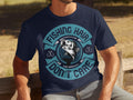 A person sits on a wooden bench wearing Garment Graphics navy blue tee, featuring a fish and the phrase Fishing Hair Dont Care in bold letters with anchor designs.