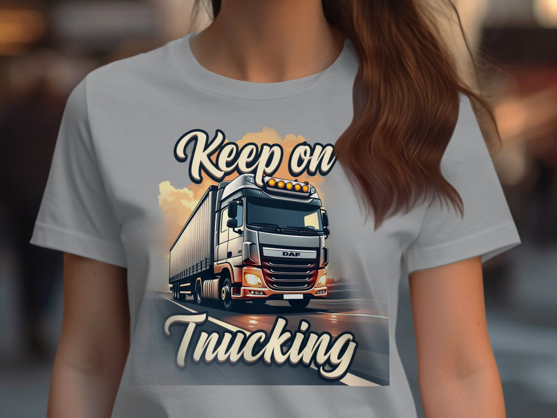 Garment Graphics white T-shirt with a truck graphic and Keep On Trucking text is displayed on wood, surrounded by striped gift boxes, pine cones, a snowflake, candy cane, and sweater.