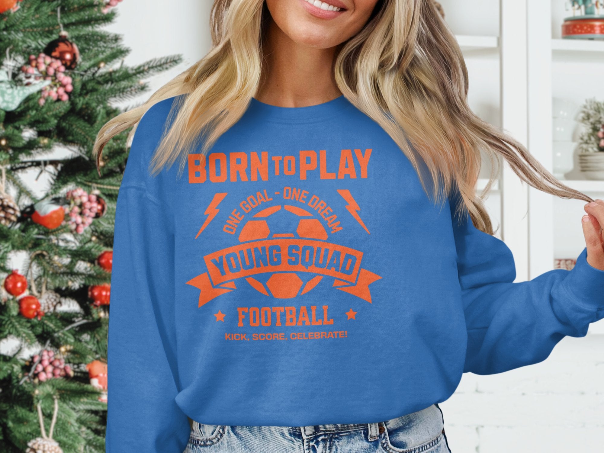 A person in a Garment Graphics blue football sweatshirt with orange text, Born to Play, One Goal - One Dream, Young Squad Football, Kick, Score, Celebrate! poses indoors. Unisex sizing fits all. A decorated Christmas tree brings festive cheer in the background.