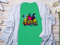 Embrace street art with Garment Graphics green T-shirt featuring I Love Graffiti in colorful graffiti style and a heart with spray cans and splatters. Displayed on a light wooden surface, its surrounded by a gray cardigan, white flowers, shoes, and jewelry.