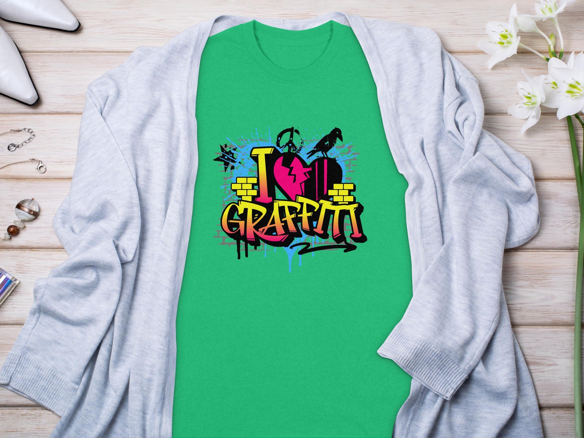 Embrace street art with Garment Graphics green T-shirt featuring I Love Graffiti in colorful graffiti style and a heart with spray cans and splatters. Displayed on a light wooden surface, its surrounded by a gray cardigan, white flowers, shoes, and jewelry.